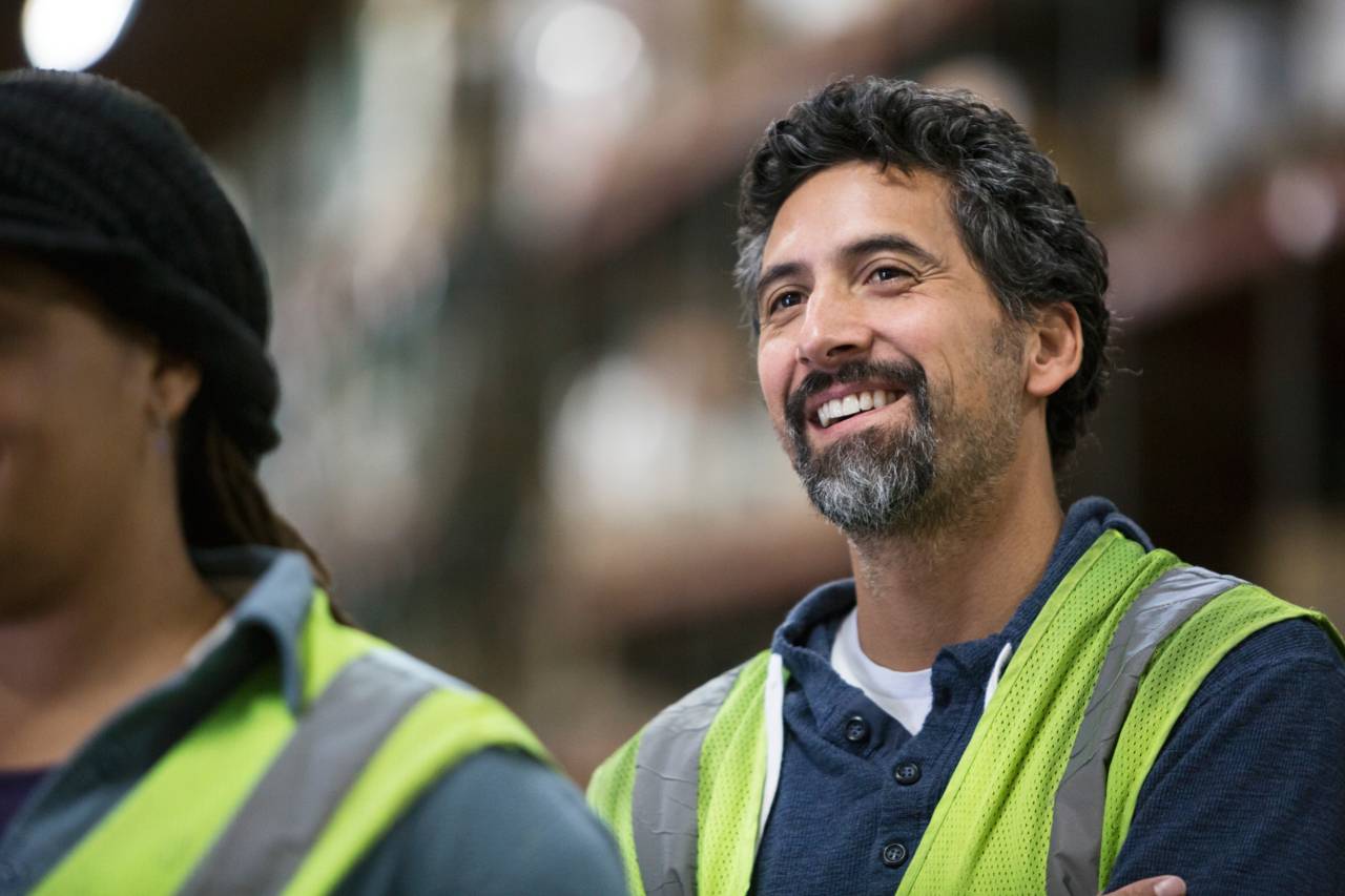 Two people wearing safety vests are smiling inside what appears to be a warehouse. Shelves and boxes are visible in the blurred background.